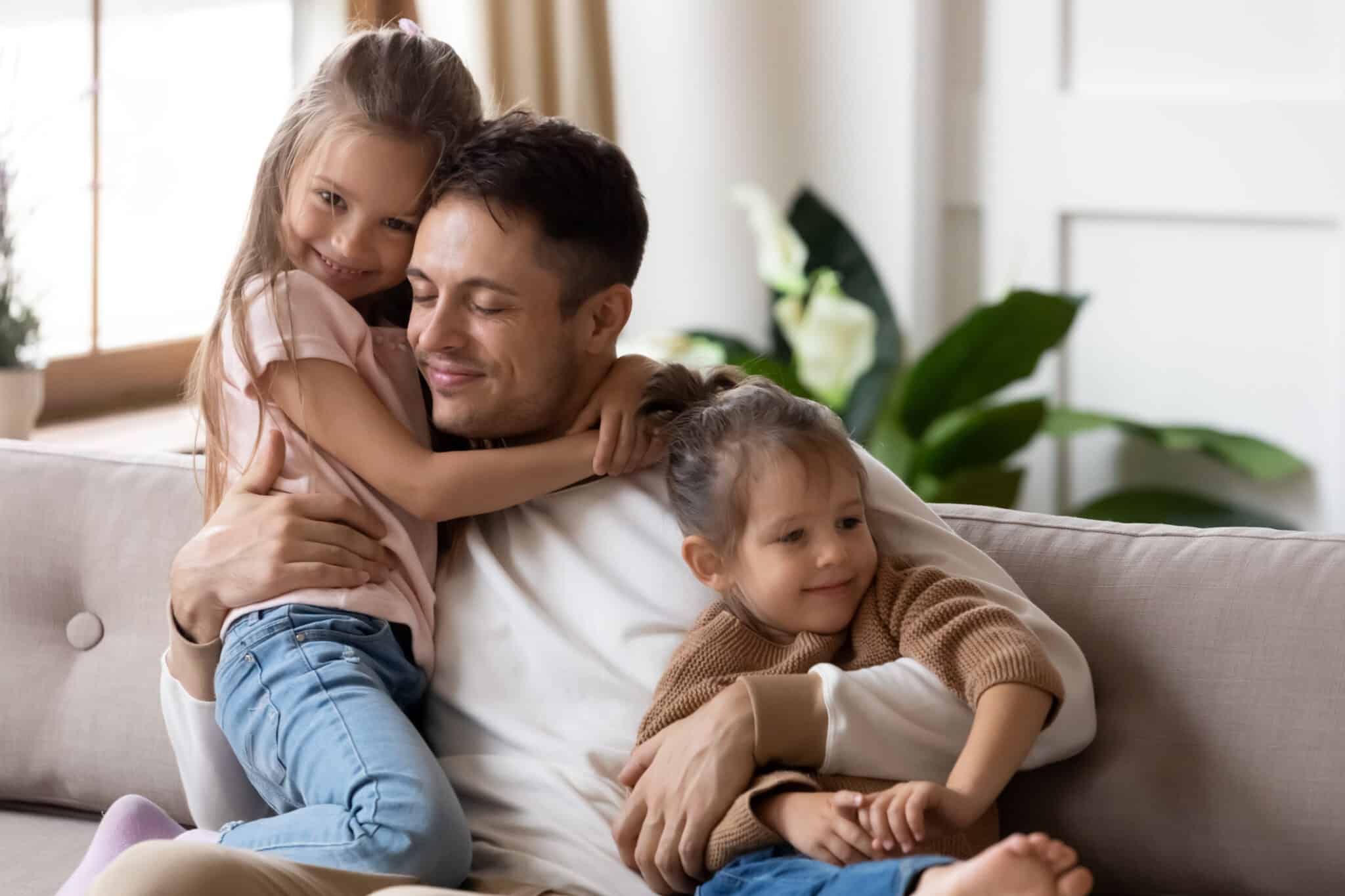 Dad hug two cute daughters resting together on couch, Whole Home Humidifiers.